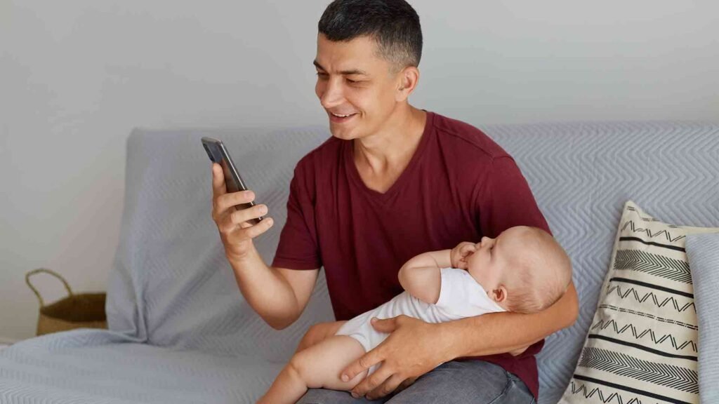 portrait-young-adult-caucasian-man-wearing-maroon-casual-style-t-shirt-with-cute-child-white-attire-father-holding-his-baby-while-using-his-phone-while-sitting-sofa-light-room