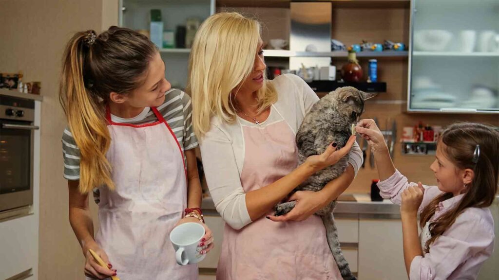 blonde-female-holds-cat-two-teenage-girl-cooking-kitchen